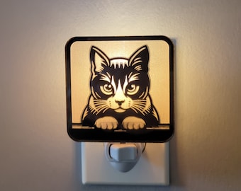 May include: A black and white cat nightlight with a white plug-in base. The cat is looking directly at the viewer with a serious expression.