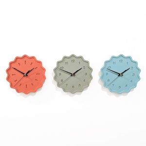 Puede incluir: Tres relojes de pared en coral, gris verdoso y azul claro. Cada reloj tiene un borde festoneado y manecillas negras. Los números están en una fuente simple y moderna. Los relojes son redondos y parecen ser de plástico.