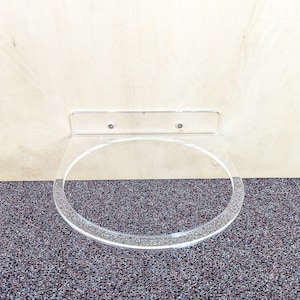 May include: A clear acrylic wall-mounted shelf for holding a round object, such as a plate or bowl. The shelf is attached to the wall with two screws.