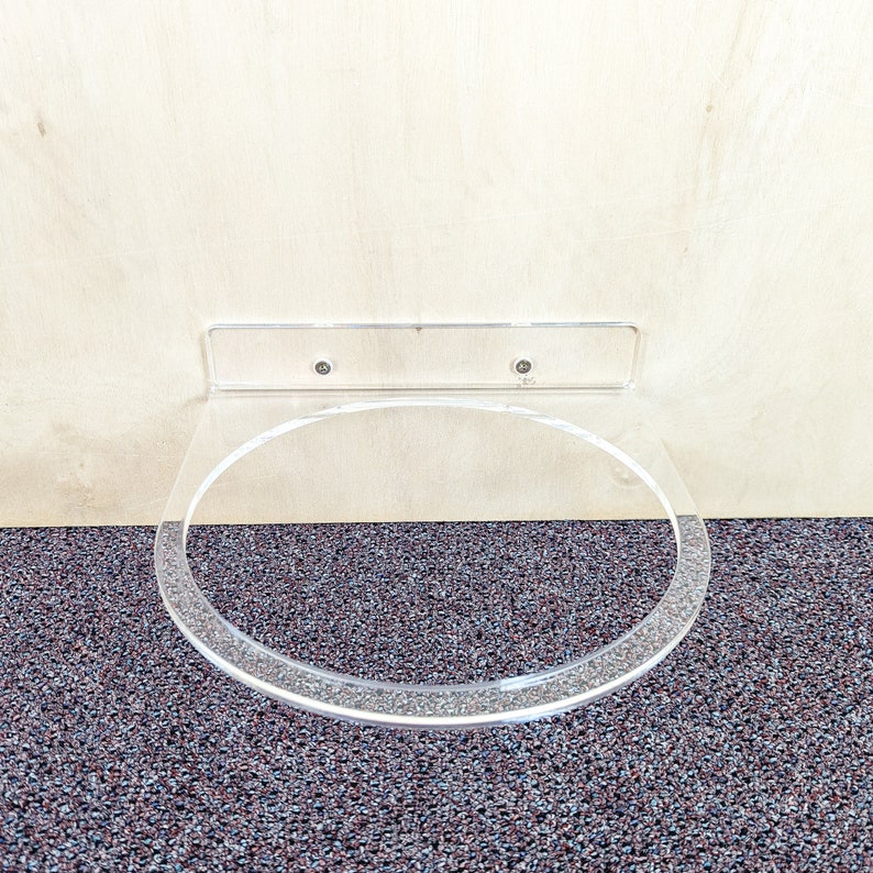 May include: A clear acrylic wall-mounted shelf for holding a round object, such as a plate or bowl. The shelf is attached to the wall with two screws.