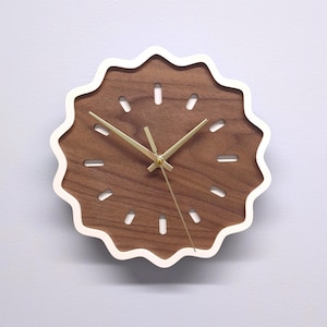 Könnte beinhalten: Eine runde Wanduhr mit einem dunkelbraunen Holzgesicht und einem weißen, wellenförmigen Rand. Die Uhr hat goldfarbene Zeiger und kleine, längliche Aussparungen um das Zifferblatt. Die Uhr hat einen Durchmesser von etwa 25 cm.