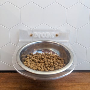 May include: A clear plastic pet food bowl holder with the word "NOM" printed on it. The holder is mounted to a white hexagon tile wall. A stainless steel bowl filled with brown pet food is attached to the holder.