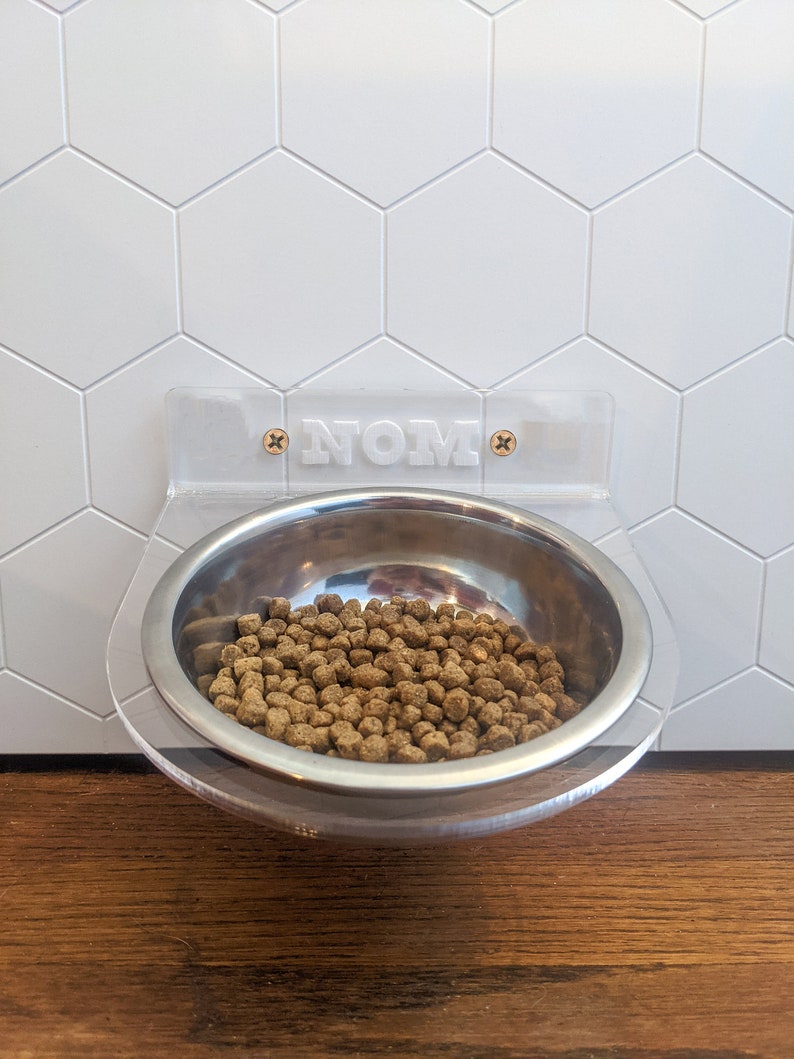 May include: A clear plastic pet food bowl holder with the word "NOM" printed on it. The holder is mounted to a white hexagon tile wall. A stainless steel bowl filled with brown pet food is attached to the holder.