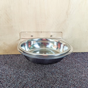 May include: A stainless steel pet bowl mounted to a wall with a clear plastic bracket. The bowl is empty and sits on a dark gray carpet.
