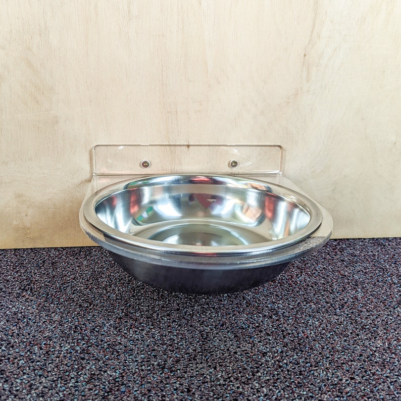 May include: A stainless steel pet bowl mounted to a wall with a clear plastic bracket. The bowl is empty and sits on a dark gray carpet.