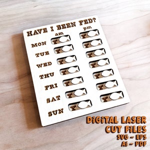 May include: A white wooden board with the text "HAVE I BEEN FED?" and a grid of 14 fish-shaped cutouts for tracking daily feeding times. The board is labeled with the days of the week and "am" and "pm". The text "DIGITAL LASER CUT FILES SVG - EPS Ai - PDF" is printed below the board.