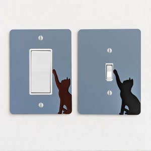 May include: Two light switch covers with a blue background and a silhouette of a cat reaching for the light switch. The switch on the left is off and the switch on the right is on.