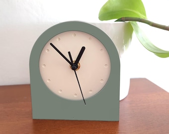 May include: A sage green, arched desk clock with a cream-colored face. The clock features black hands and small dot hour markers. The clock is set on a wooden surface, with a white ceramic pot and green plant in the background.
