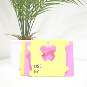 Puede incluir: Una tarjeta amarilla con un recorte en forma de corazón y el texto "USE BY" impreso en ella. La tarjeta está encima de una tarjeta rosa.