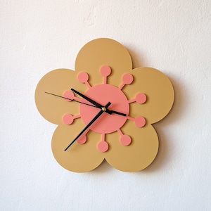 May include: A flower-shaped clock with a pink center and black hands. The clock has a light brown background and is mounted on a white wall.