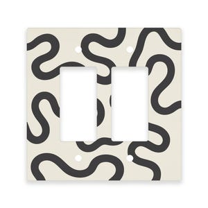 May include: A decorative light switch plate with two openings. The plate has a cream-colored background with a repeating pattern of black, wavy lines. The design is modern and abstract, suitable for home decor.