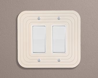Op de afbeelding: Een witte dubbele lichtschakelaar met een crèmekleurige omlijsting. De omlijsting heeft een decoratief, concentrisch cirkelontwerp.