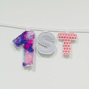 May include: A fabric banner spelling out HAPPY 1ST BIRTHDAY, featuring individual stuffed letters in various patterns and colors including pink, purple, blue, and white. Each letter is suspended on a thin white string against a plain background.