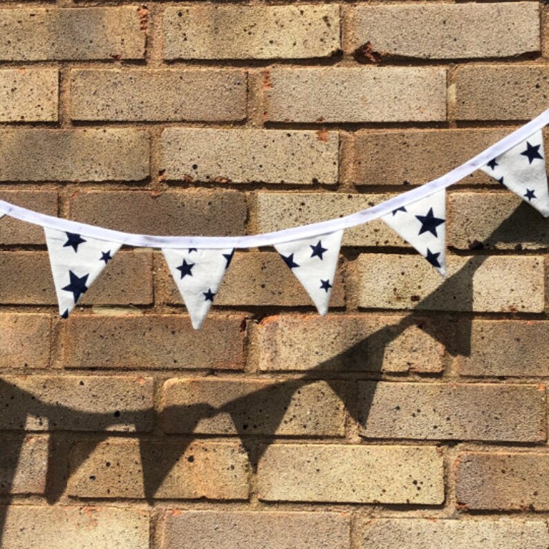May include: A white bunting with navy blue stars hanging against a brick wall. The bunting is made of fabric and has a white string running through it.
