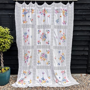 Può includere: Tenda floreale bianca con bordo in pizzo. La tenda presenta un design patchwork con bouquet floreali colorati in quadrati. Il bordo in pizzo e il bordo superiore aggiungono un tocco delicato. La tenda è appesa a un'asta.
