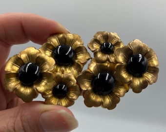 Puede incluir: Un broche de metal dorado con cinco elementos en forma de flor dispuestos en racimo. Cada flor tiene pétalos texturizados y radiantes con una piedra cabujón negra, lisa, redonda y brillante, engastada en el centro.
