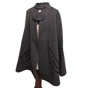 May include: A dark gray wool cape with black trim and decorative buttons. The cape features a high collar and a flowing design, suitable for outerwear. The black trim adds a contrasting detail to the cape's overall aesthetic.