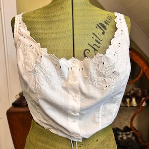 May include: A white sleeveless top featuring a scalloped neckline with eyelet embroidery and floral patterns. The garment has a central vertical button placket with small white buttons and vertical seams, displayed on a green dress form.