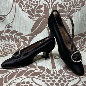 May include: A pair of black satin vintage shoes with small heels. Each shoe features a decorative embellishment with a cluster of fabric and a ring of clear rhinestones. The shoes have a strap that goes around the ankle.