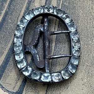 May include: An oval-shaped metal buckle featuring a dark, weathered finish and a central prong. The outer frame is encrusted with a series of clear, faceted stones arranged in a continuous border around the perimeter of the buckle structure.