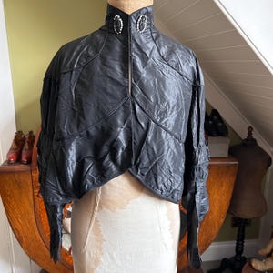 May include: A black cropped jacket with a high collar, featuring two oval-shaped decorative brooches with clear crystals and black centers at the neckline. The jacket has long sleeves with gathered detailing and a front hook-and-eye closure.