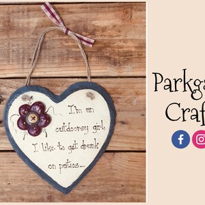 May include: A heart-shaped slate sign with a painted flower and the text "I'm an outdoorsy girl, I like to get drunk on patios..." hanging on a wooden background.