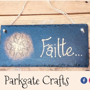 Può includere: Un cartello in ardesia nera con un disegno di nodo celtico dorato e la parola "Fáilte" scritta in lettere dorate. Il cartello è appeso a una corda e ha uno sfondo in legno rustico.