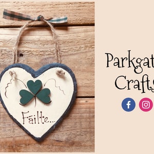Può includere: Un cartello in ardesia a forma di cuore bianco con un trifoglio verde e la parola "Fáilte..." scritta in nero. Il cartello è appeso a uno spago di iuta con un nastro a quadri legato in cima.