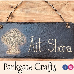Può includere: Un cartello in ardesia nera con un disegno di albero a nodo celtico dorato e il testo "Àit Shona." Il cartello è appeso a una corda di spago.