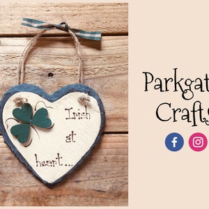 Può includere: Un cartello in ardesia a forma di cuore con un trifoglio verde e il testo "Irish at heart..." appeso su uno sfondo di legno. Il cartello è decorato con un nastro verde e bianco.
