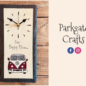 May include: A black slate clock with a white background and a red and white vintage camper van illustration. The clock face has black hands and a gold center. The text "Our Happy Place..." is written below the clock face.