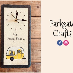 May include: A black slate clock with a white background and a yellow camper van illustration. The clock face has black hands and a gold center. The text "Our Happy Place..." is written in black on the white background.