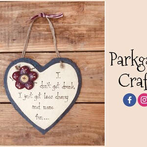 May include: A heart-shaped slate sign with a red flower and the text "I don't get drunk, I just get less classy and more fun..." hanging on a wooden background.