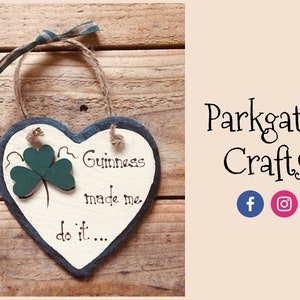 Può includere: Un cartello in ardesia a forma di cuore bianco con bordo nero è appeso a uno spago di iuta. Il cartello recita "Guinness made me do it..." con un trifoglio verde dipinto sul lato sinistro.