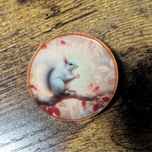 May include: A round, resin-coated pin featuring a white squirrel perched on a branch with red leaves. The squirrel is the focal point, set against a soft, light background. The pin has a brown border.