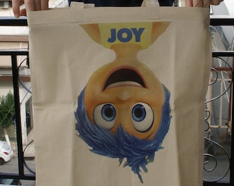 Könnte beinhalten: Eine beige Einkaufstasche mit einer Comicfigur von Freude aus dem Film Inside Out. Freude ist auf dem Kopf stehend und hat blaues Haar und große blaue Augen. Der Text "FREUDE" ist auf ihrem Shirt gedruckt.