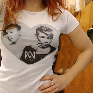 May include: White t-shirt with a heart-shaped design featuring two boys with the text "Marcus & Martinus" and the logo "MM" in black.