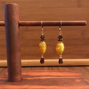 May include: A pair of gold and burgundy earrings with a teardrop-shaped glass bead and a smaller burgundy bead. The earrings are hanging from a wooden stand.