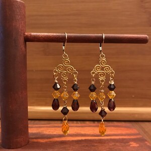 May include: Gold dangle earrings with a geometric design and three tiers of dangling crystals. The crystals are a gradient of dark purple, amber, and gold.