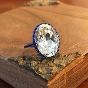 Könnte beinhalten: Ein Ring mit einem großen, klaren ovalen Kristall, der in einer blauen Metallfassung sitzt. Der Ring liegt auf einer Holzoberfläche.
