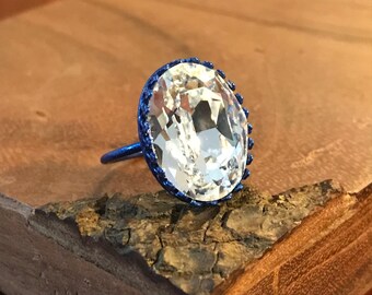 May include: A ring with a large, clear oval-shaped crystal set in a blue metal bezel. The ring is on a wooden surface.