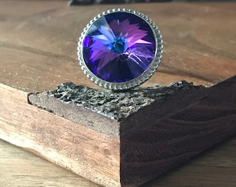 May include: A round, purple, iridescent crystal with a silver bezel setting. The crystal is set on a wooden surface.