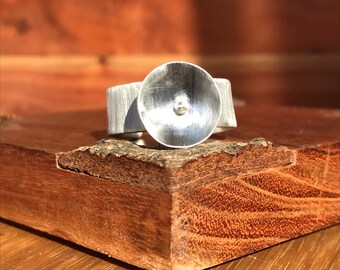 May include: A silver ring with a textured band and a large, circular, brushed metal top. A small, round silver detail is centered on the top. The ring is displayed on a wooden surface.