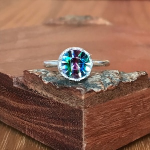 May include: A silver ring with a large, round, iridescent gemstone. The gemstone displays a spectrum of colors, including blues, greens, and purples. The ring is set on a rustic wooden surface, showcasing the ring's design and colors.