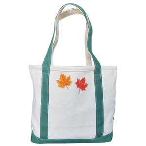 May include: A white canvas tote bag with green trim and handles. Two embroidered maple leaves, one orange and one red, are centered on the front. The bag is designed for carrying items.