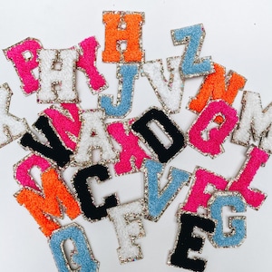 May include: A collection of colorful felt letters with gold glitter trim. The letters are in various colors, including pink, blue, orange, black, and white. The letters are arranged in a random order.