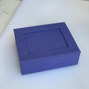 May include: A rectangular purple box with a flat lid featuring a recessed central panel. The exterior of the box has a textured fabric-like finish, and the lid is designed to fit securely over the base, creating a clean and uniform appearance.