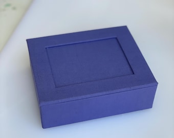 May include: A rectangular purple box with a flat lid featuring a recessed central panel. The exterior of the box has a textured fabric-like finish, and the lid is designed to fit securely over the base, creating a clean and uniform appearance.