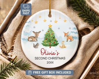 May include: A round white ceramic ornament featuring a watercolor illustration of a green Christmas tree, a deer, a fox, a rabbit, and a squirrel. The text reads Olivia's SECOND CHRISTMAS 20XX, accented by hanging gold stars and snowflakes.