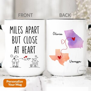 May include: A white ceramic mug with a black handle featuring two views. The front displays the text MILES APART BUT CLOSE AT HEART with line drawings of two houses and a paper airplane. The back shows watercolor maps of Georgia and Texas.